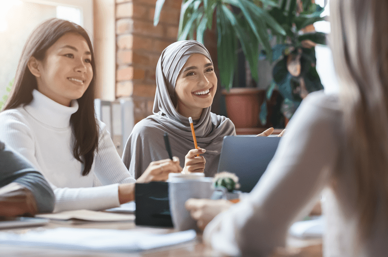 Two women, one wearing hijab and one not, sit at a table discussing business